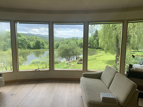 view out of a home with five big windows overlooking a pond with trees and meadow surrounding it, lush, green, and peaaceful